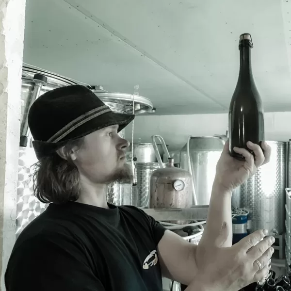 Māris Plūme apskata tikko aizkorķētu sidra pudeli. Fonā sidra mucas.